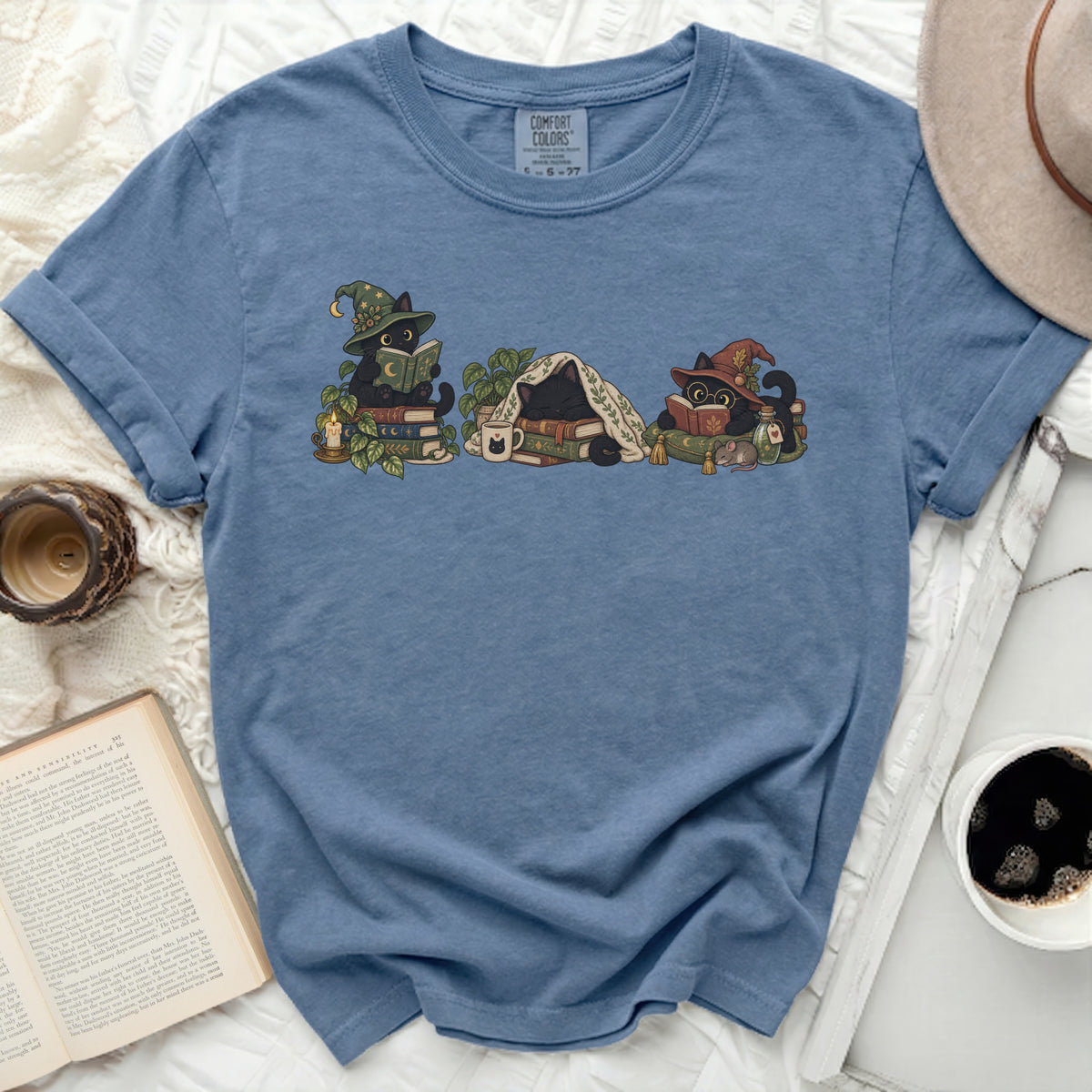 Blue t-shirt with a graphic of three black cats reading books surrounded by plants and books.