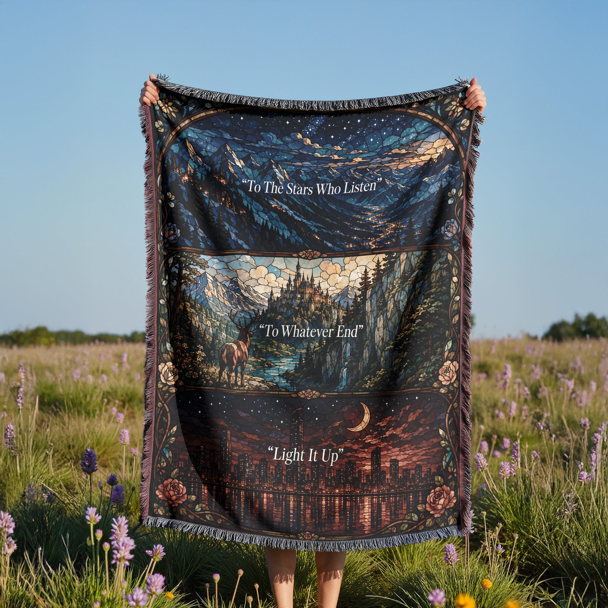 A person is holding a large, colorful blanket with a night sky and landscape design printed on it, standing in a field with purple flowers.