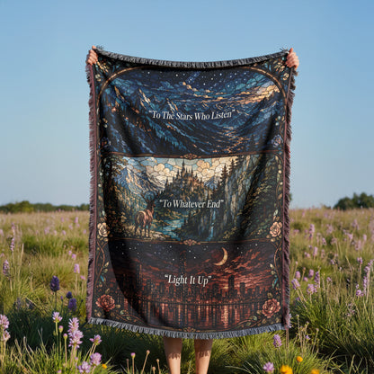 A person is holding a large, colorful blanket with a night sky and landscape design printed on it, standing in a field with purple flowers.
