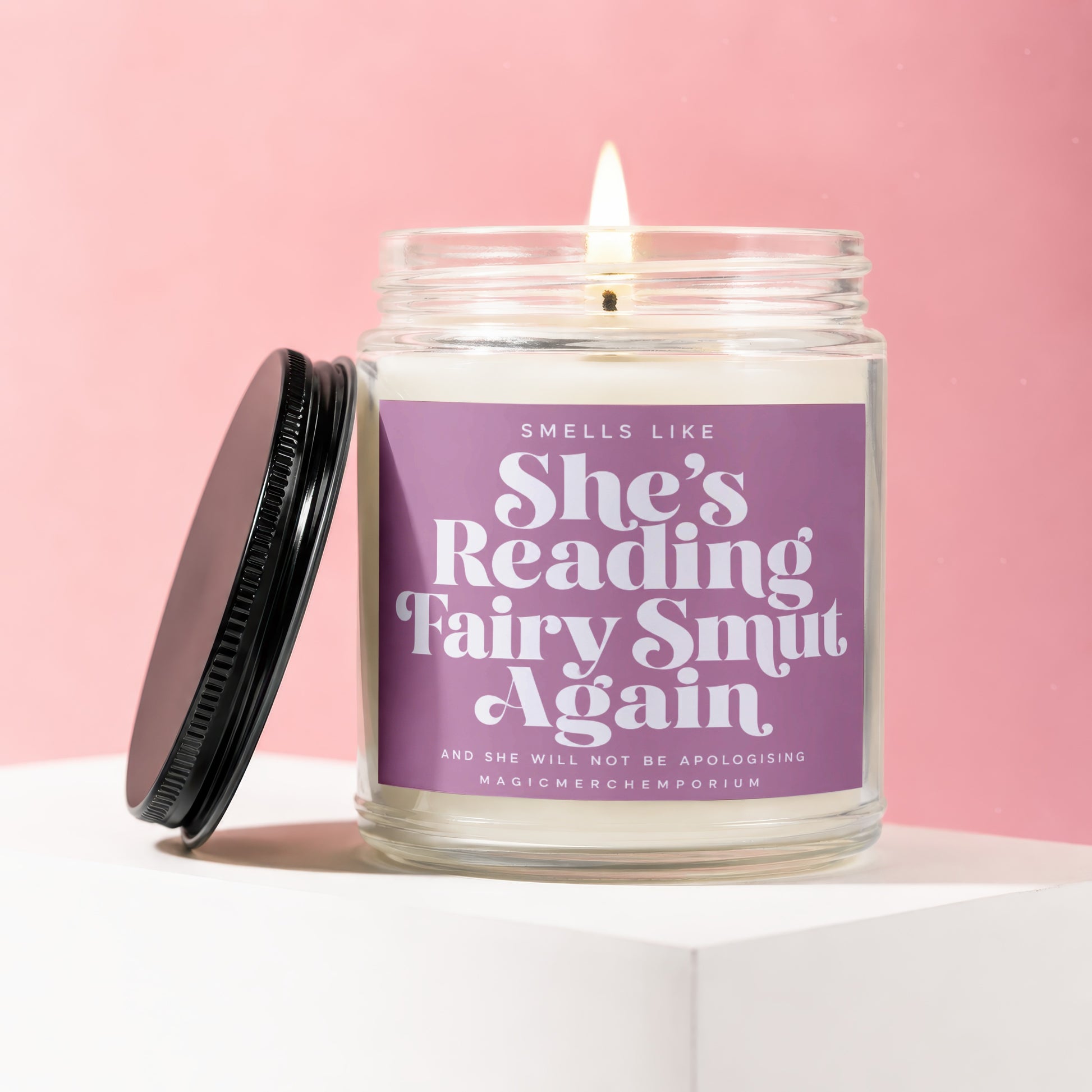 Lit candle in a glass jar with a purple label that reads "Smells Like She's Reading Fairy Smut Again".