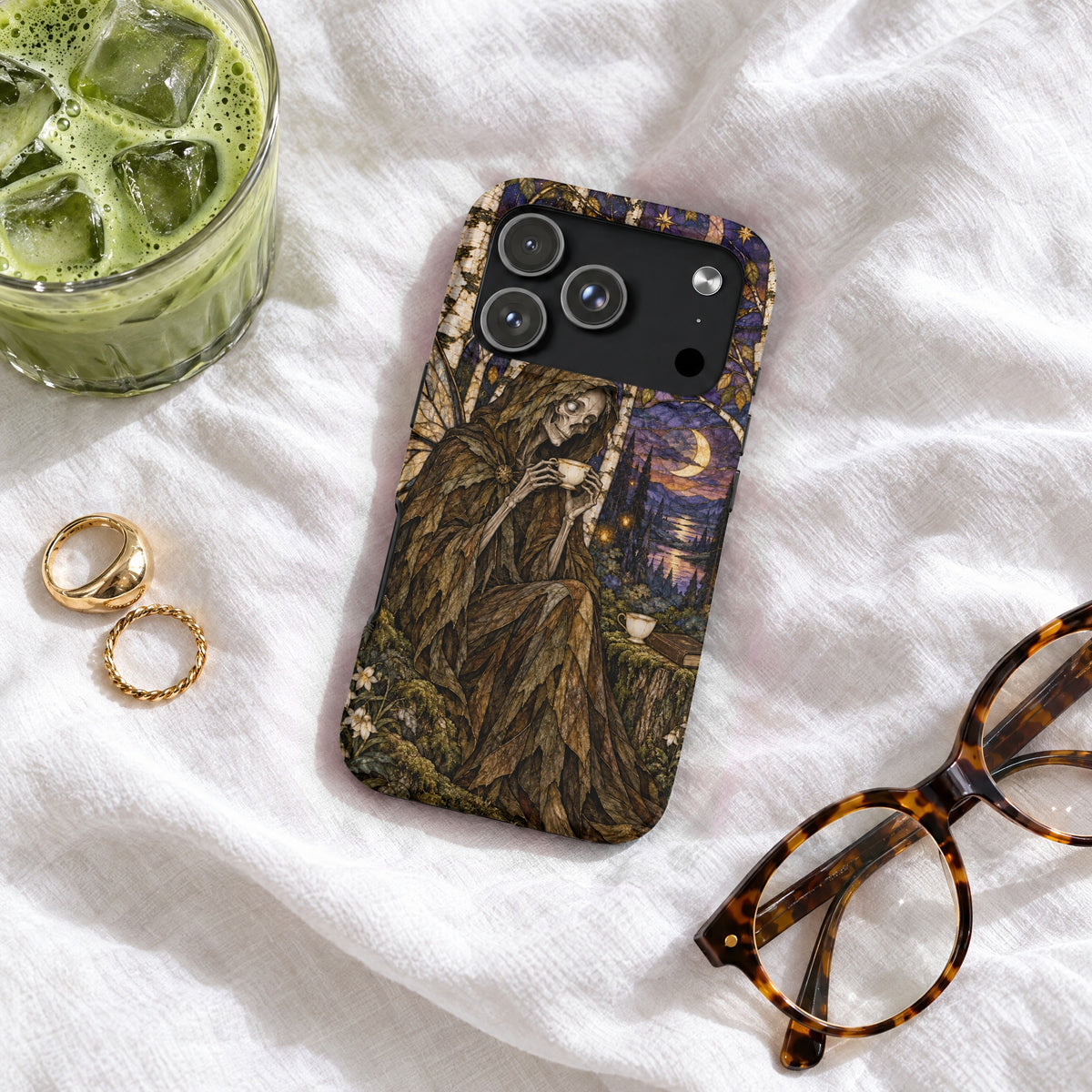 A smartphone case featuring a skeletal figure drinking tea is laid on a white fabric surface alongside glasses, rings, and iced matcha.