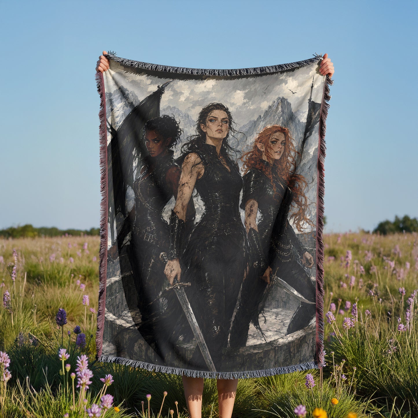 A person is holding up a large blanket with a printed image of three women on it, standing in a field with purple flowers.