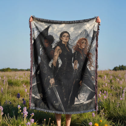 A person is holding up a large blanket with a printed image of three women on it, standing in a field with purple flowers.