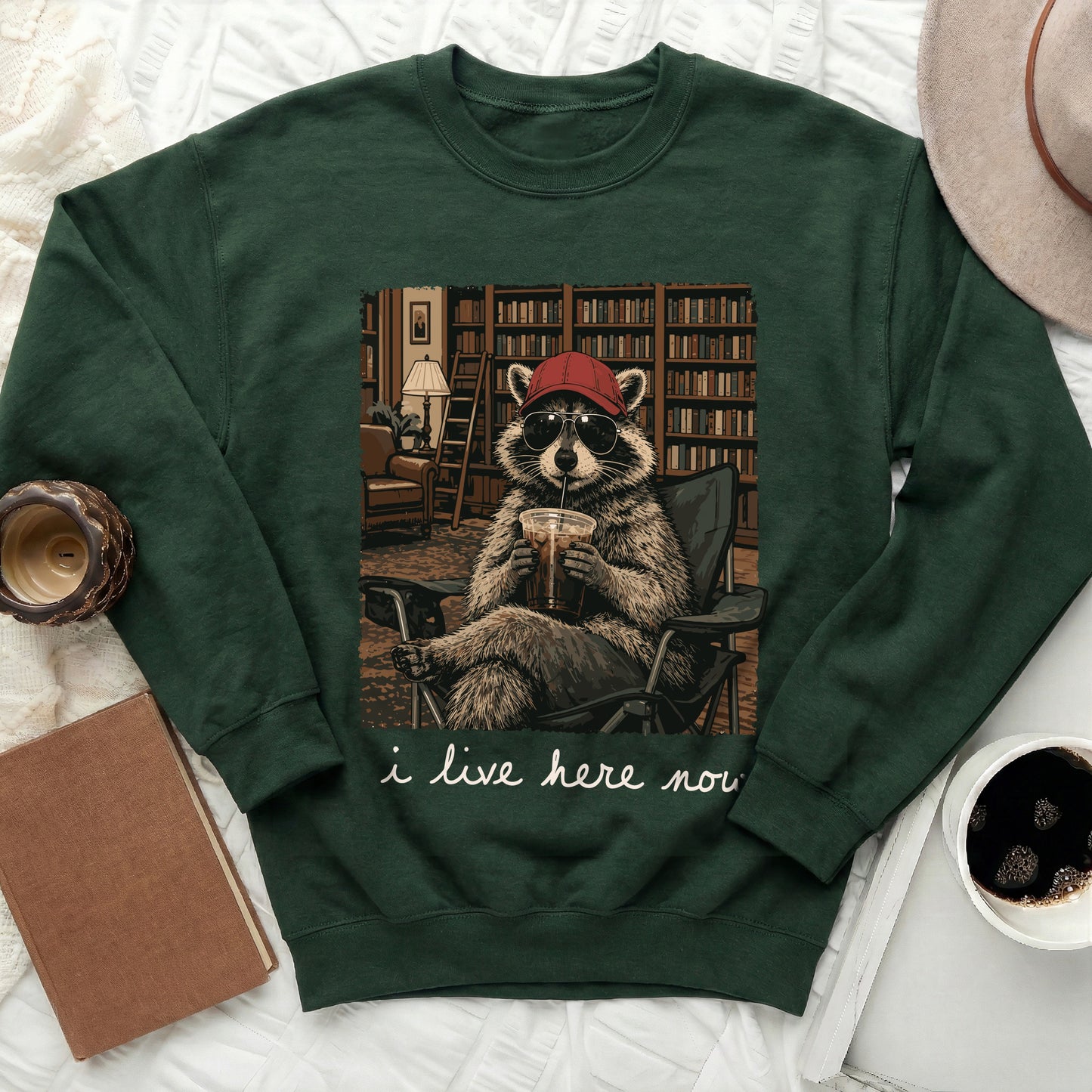Green sweatshirt with raccoon graphic in library, holding drink, wearing hat and sunglasses.