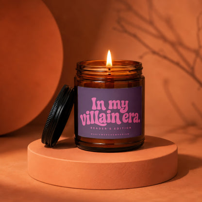 Amber candle with lit flame, purple label reading "In my villain era. READER'S EDITION MAGICMERCH.EMPORIUM".