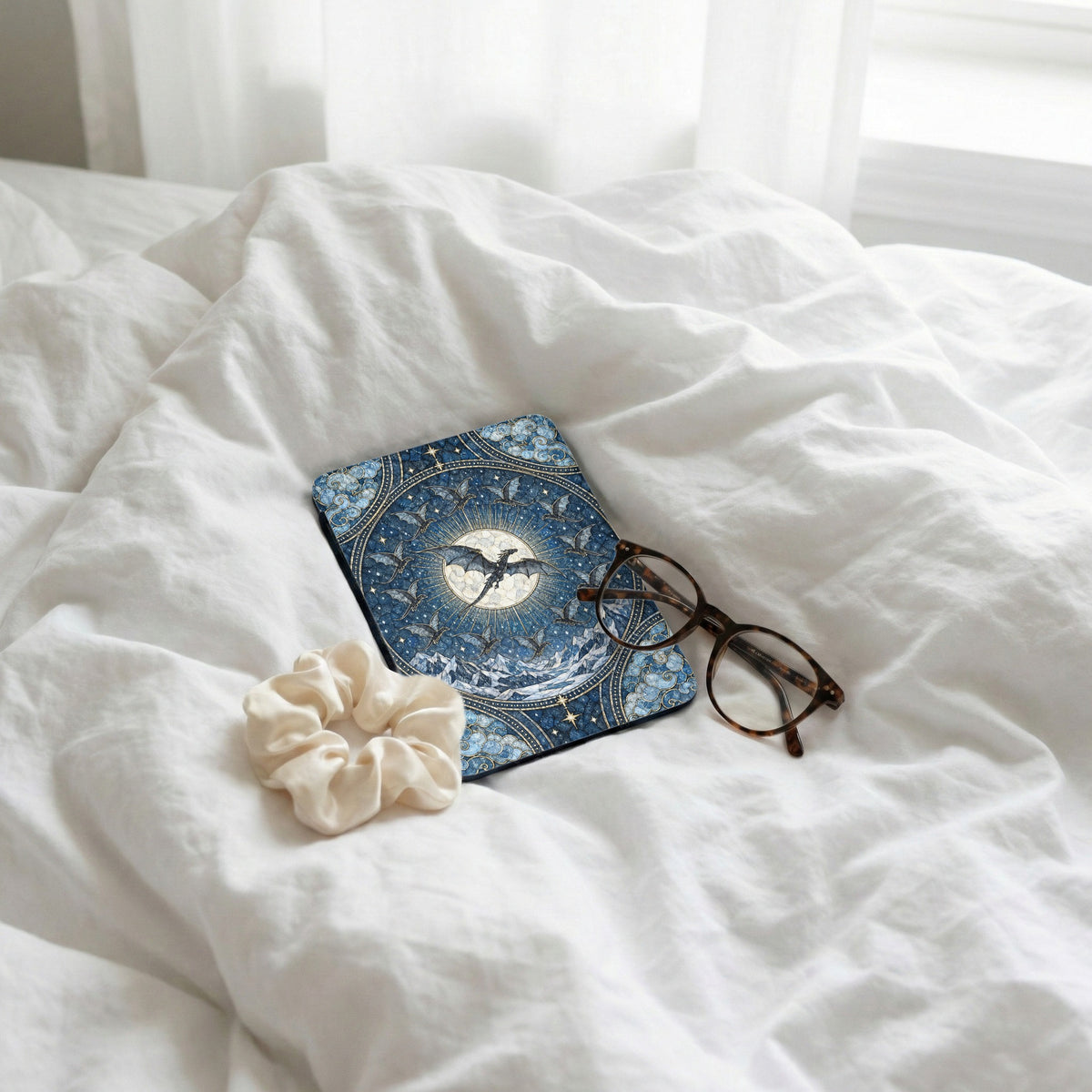 A blue fantasy-themed e-reader case lies on a white bed with glasses and a scrunchie.