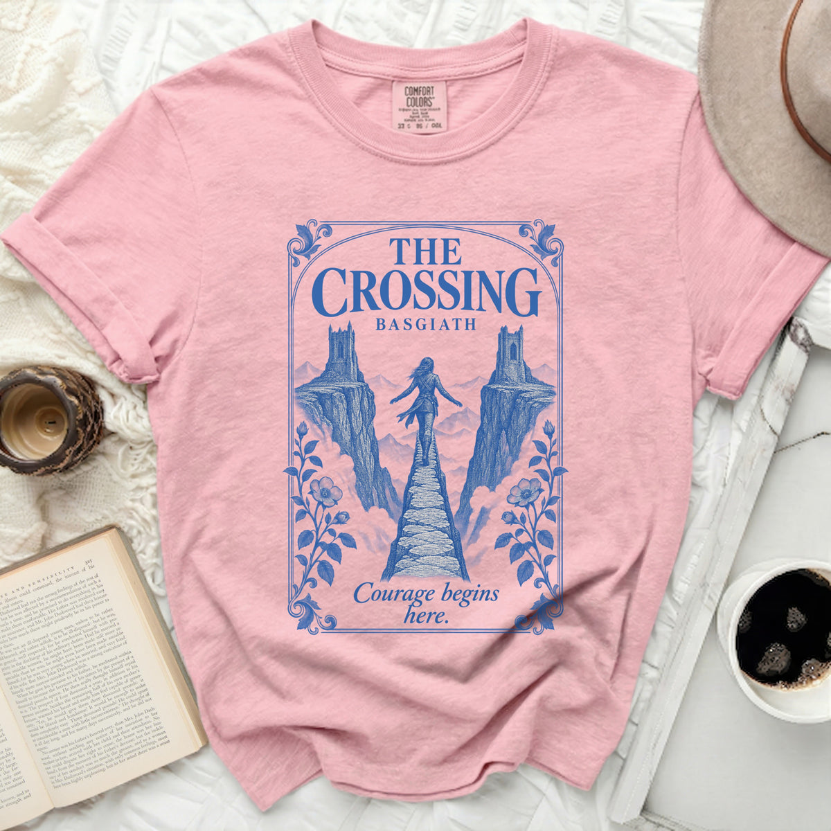 A pink crewneck t-shirt featuring a blue graphic of a person crossing a stone bridge between cliffs, with the text The Crossing Basgiath and Courage begins here.