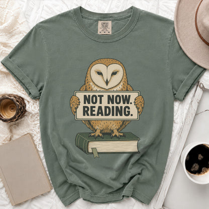 Green t-shirt with owl graphic holding "NOT NOW. READING." sign, on book.