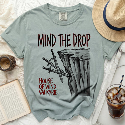 Light blue t-shirt with "MIND THE DROP" and swords on a cliff graphic.