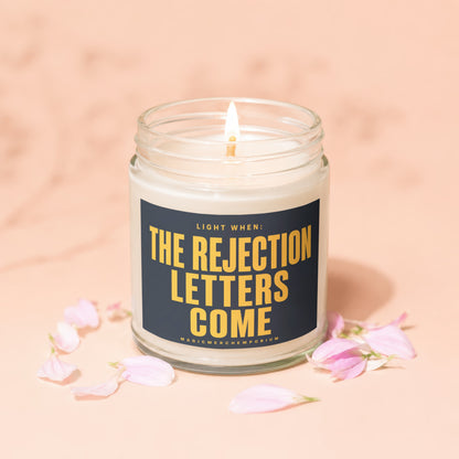 Glass jar candle with lit wick, dark label reads "Light When: The Rejection Letters Come."