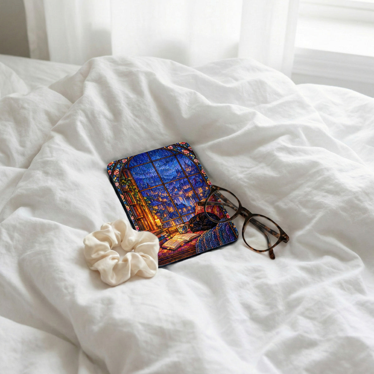 An e-reader with a colorful stained-glass window design rests on rumpled white bedding alongside tortoiseshell glasses and a white scrunchie.