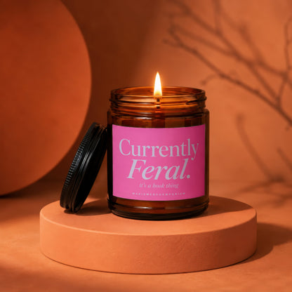 Amber glass candle with "Currently Feral. it's a book thing" text on a pink label.