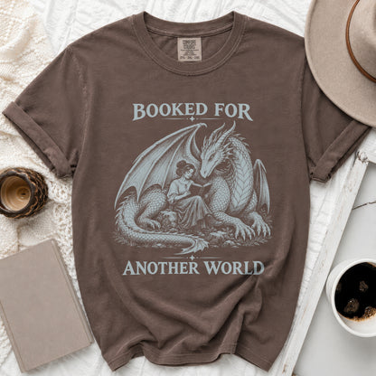 Brown t-shirt with dragon and woman reading, text "BOOKED FOR ANOTHER WORLD".