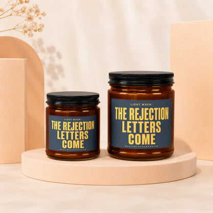 Two amber jar candles with black lids and labels reading "The Rejection Letters Come".