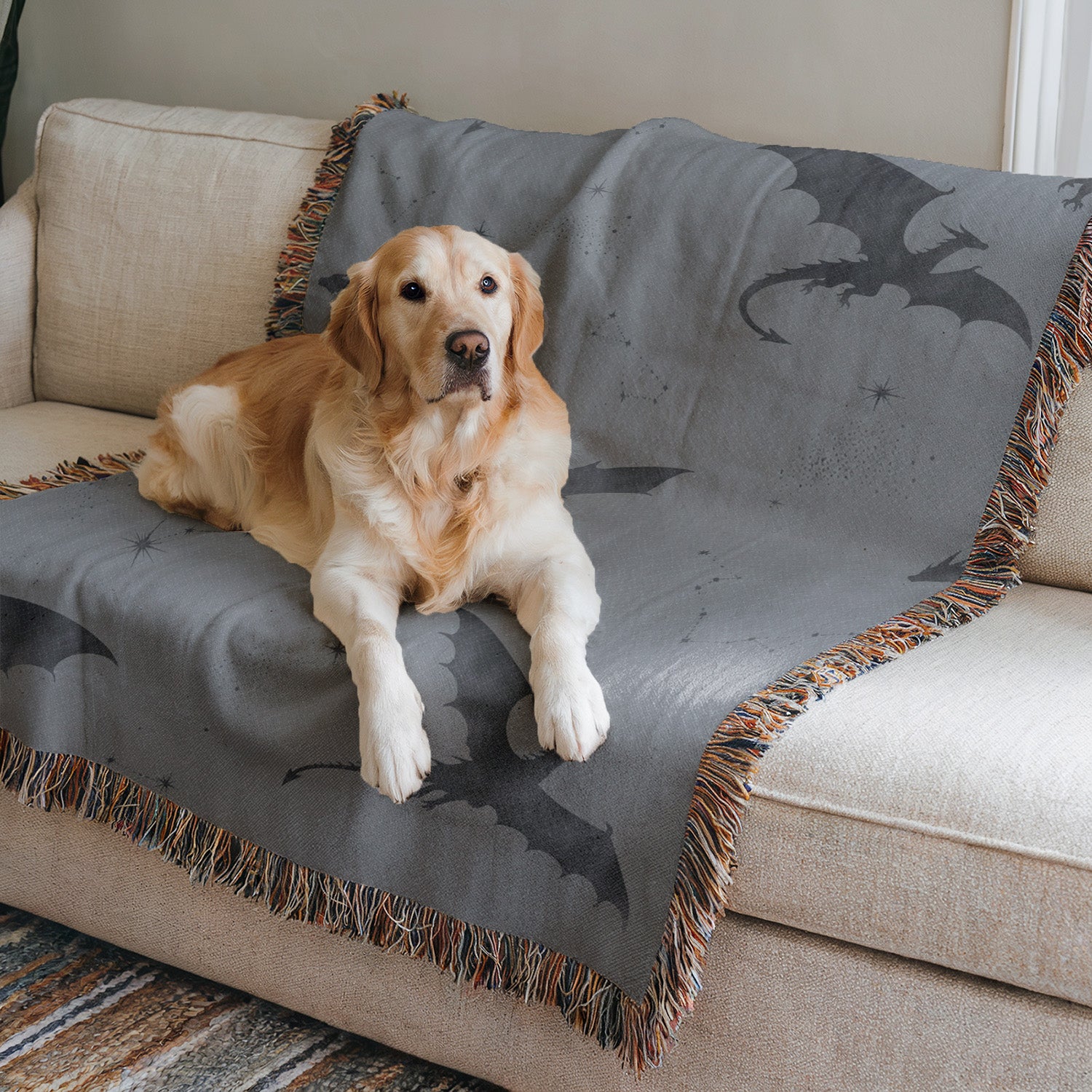 A golden retriever dog is sitting on a gray blanket with a dragon design, which is placed on a beige couch.