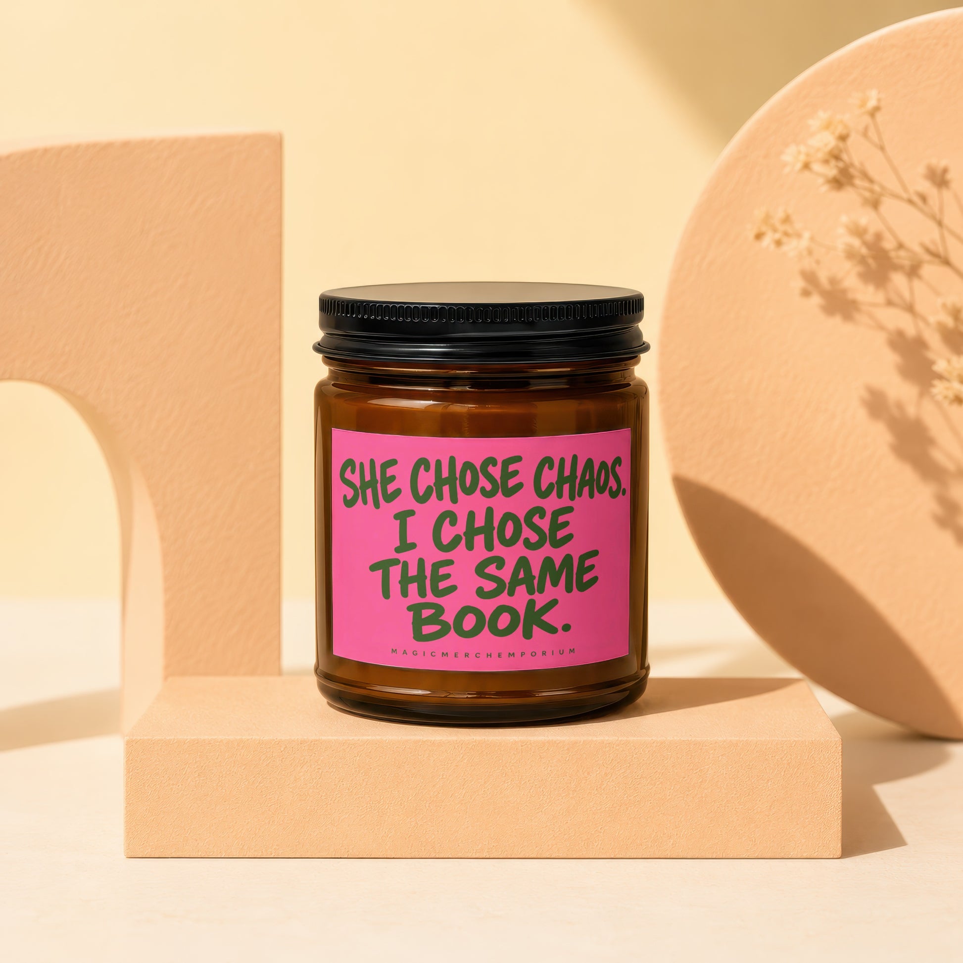 Brown candle with pink label reading "SHE CHOSE CHAOS. I CHOSE THE SAME BOOK."