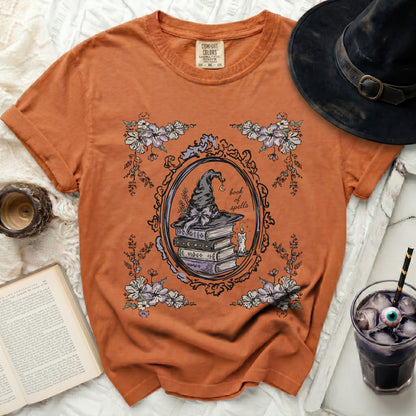 Orange t-shirt with a vintage-style graphic of a witch's hat, books, and flowers.