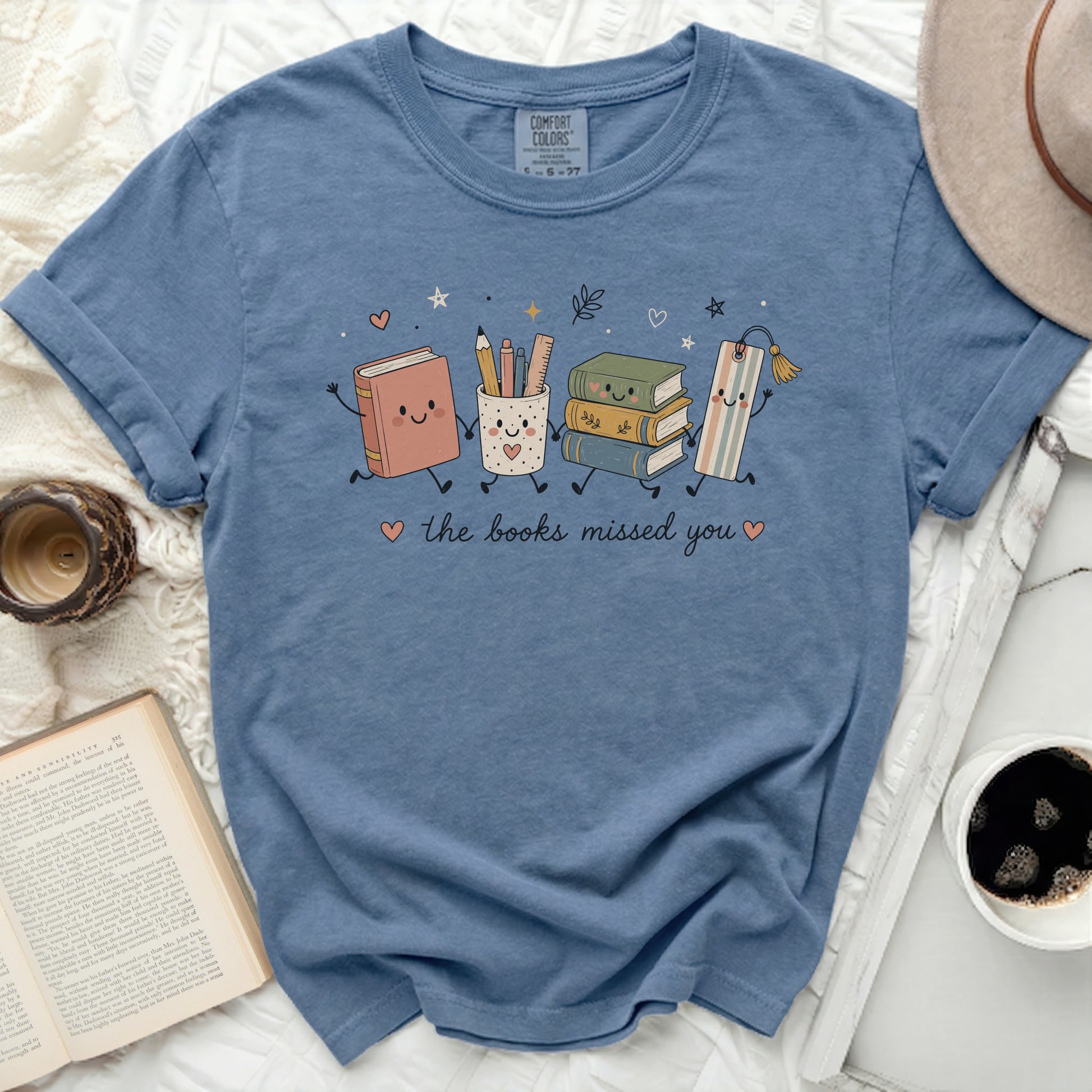 Blue t-shirt with graphic of smiling books and school supplies, "the books missed you."