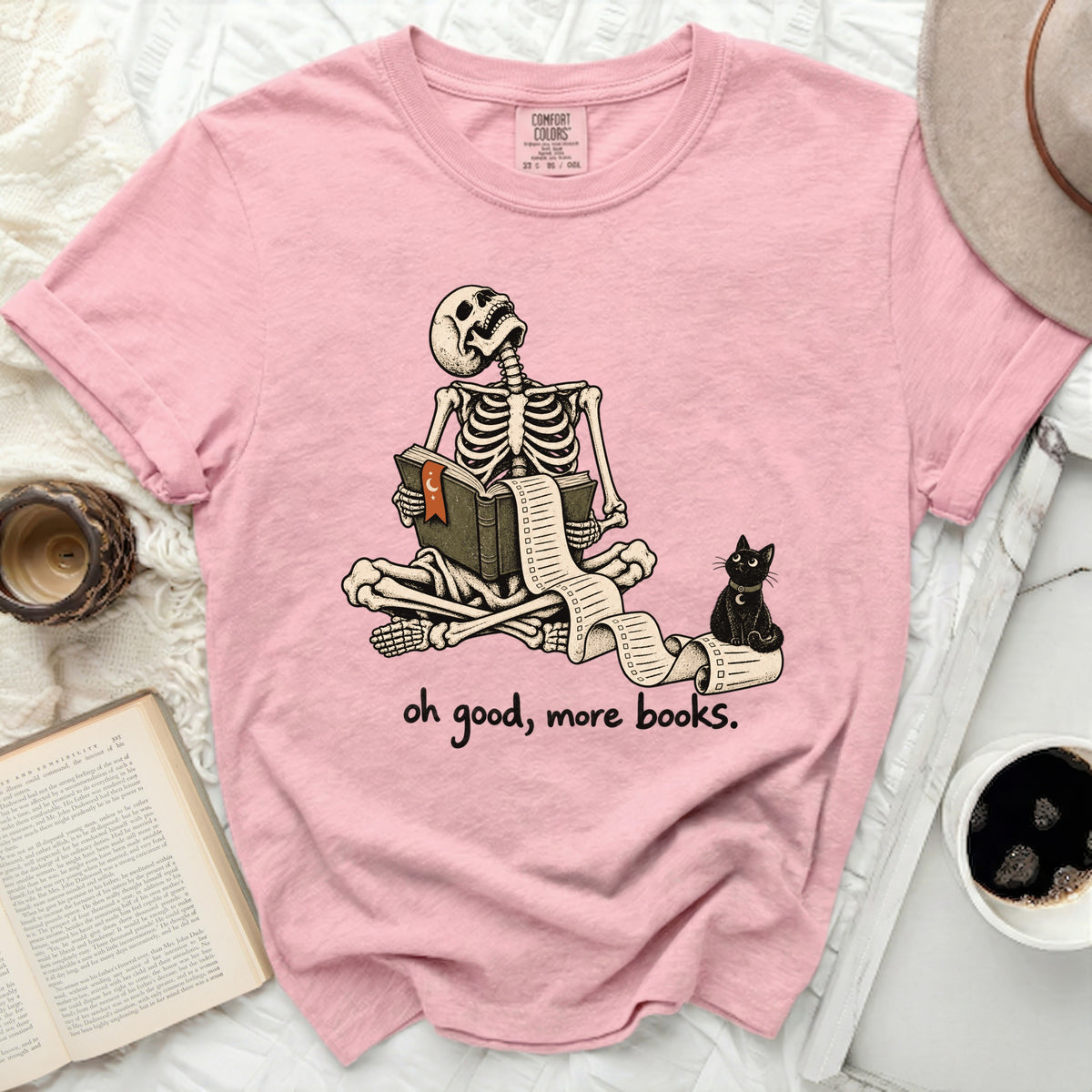 Pink t-shirt with skeleton reading a long scroll and a black cat saying "oh good, more books."