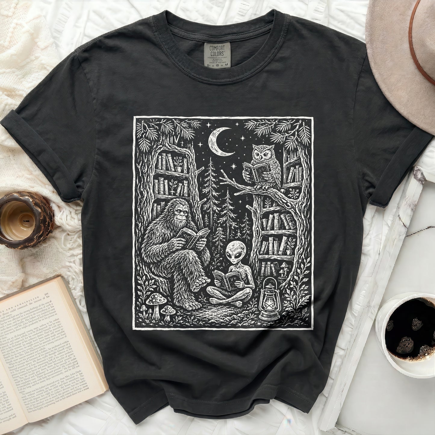 Black t-shirt with a print of Bigfoot, alien, owl reading in a forest with bookshelves under a moon.