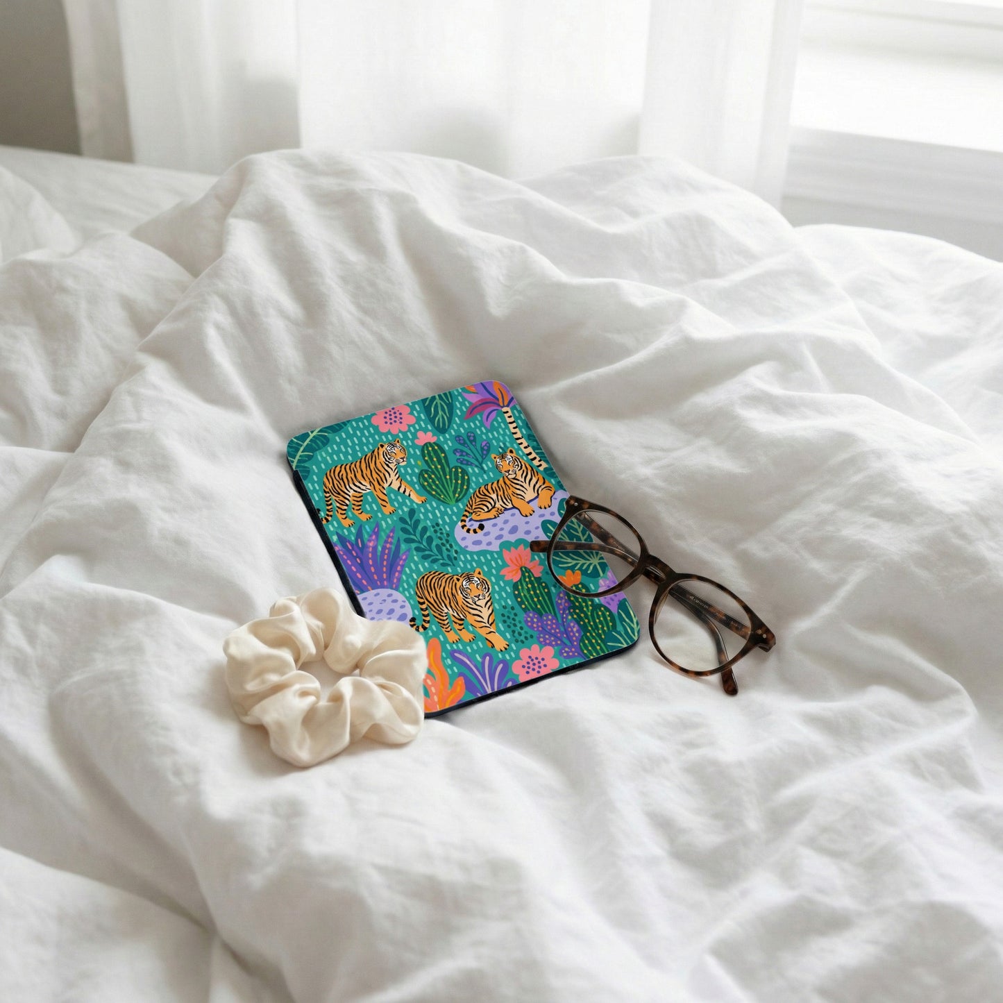 Case with tigers and plants, scrunchie, and glasses on white crumpled sheets.