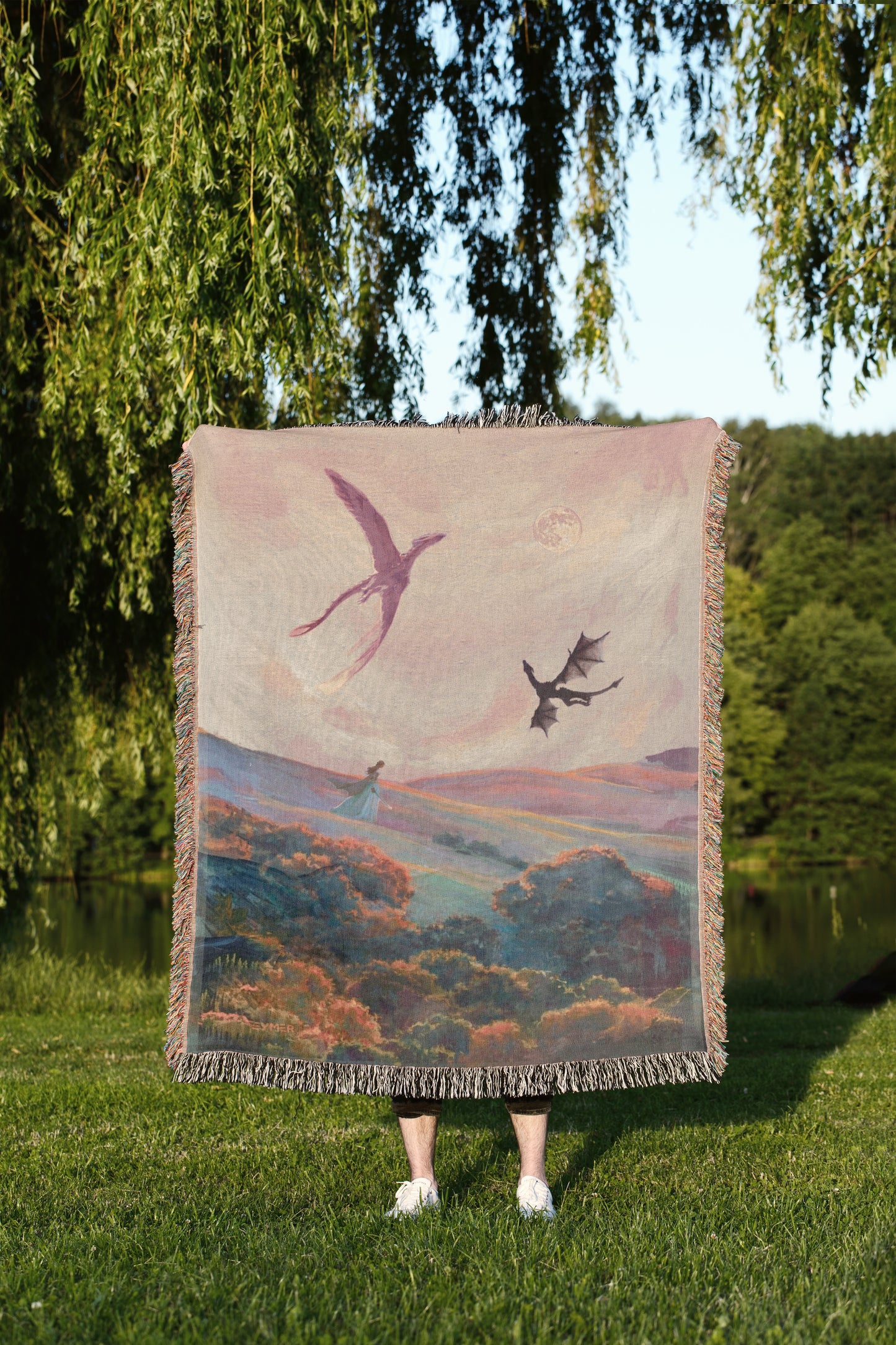 A person stands in a grassy field, holding a large woven blanket with a painting of a bird and a dragon on it.