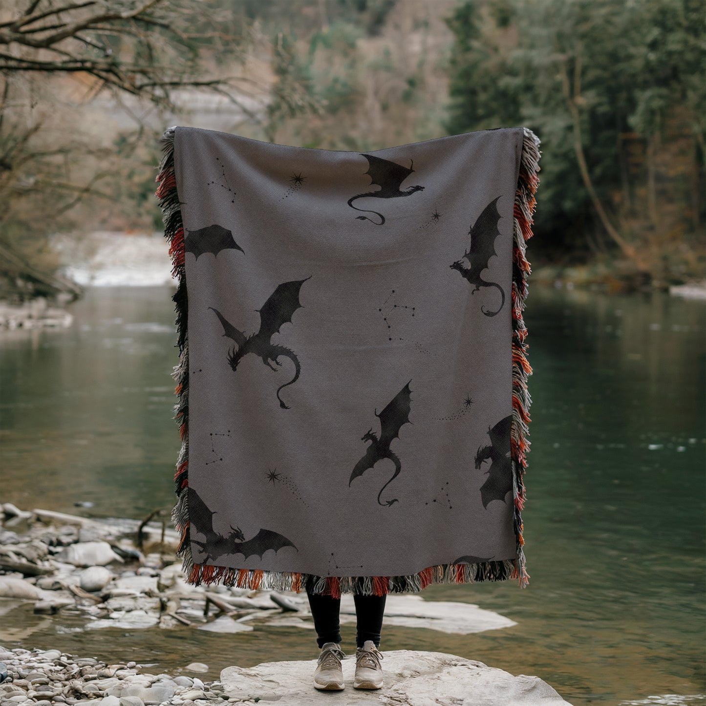 A person is standing on a rock, holding a large blanket with a dragon design on it, partially covering their face.