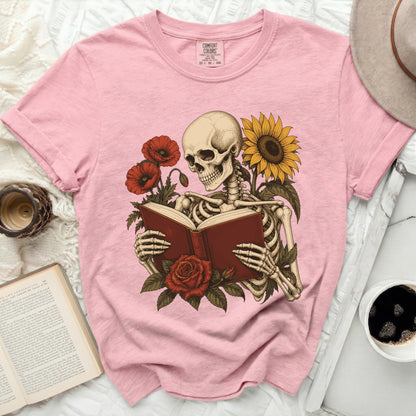 Pink t-shirt with skeleton reading a book surrounded by poppies and a sunflower.