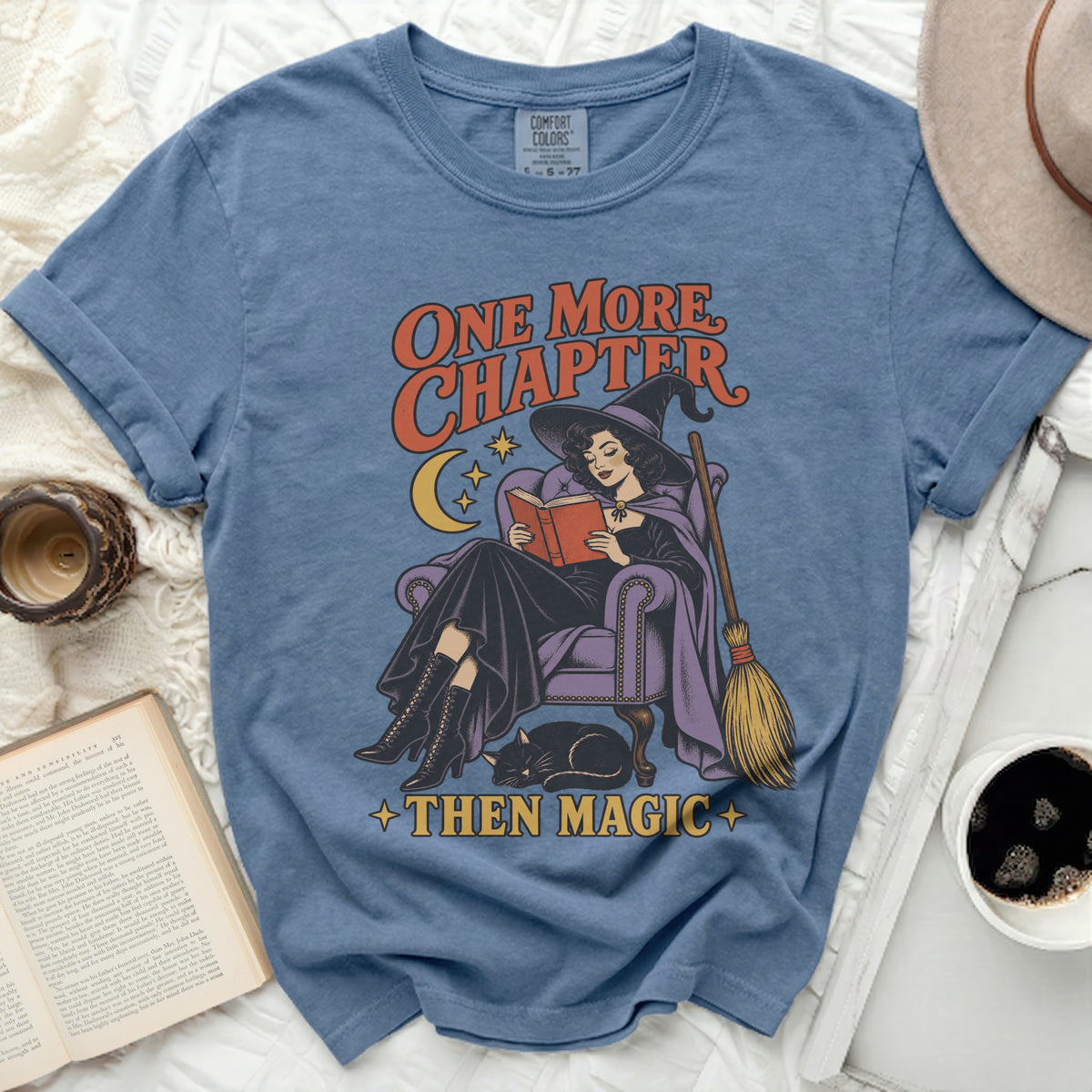 Blue t-shirt with graphic of witch reading a book, cat, broom, and text "ONE MORE CHAPTER THEN MAGIC".