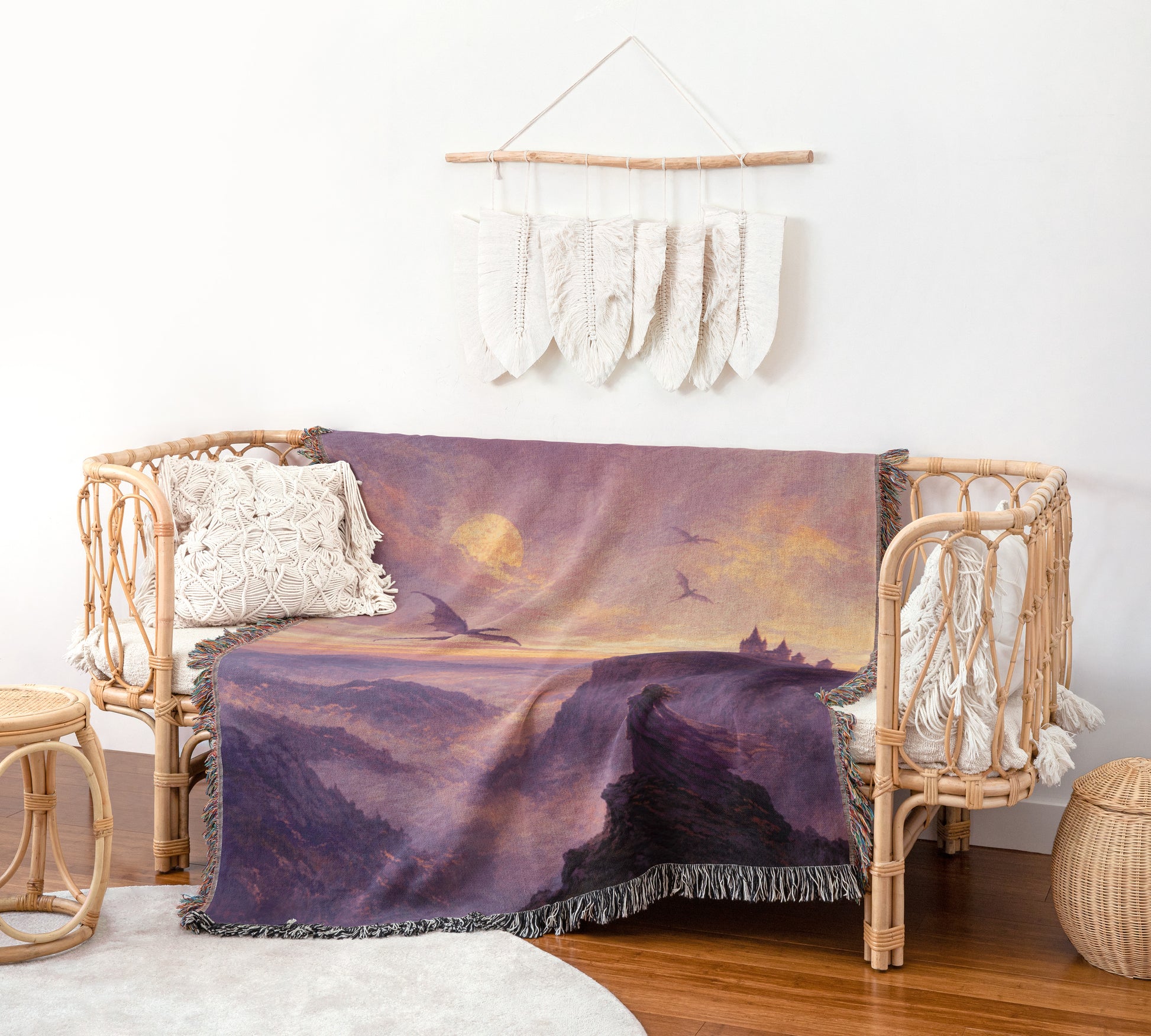 a cozy living room with a wicker bench, a woven basket, and a woven side table. The bench is adorned with a purple blanket that depicts a mountain landscape, and a hanging wall decoration with white feathers.