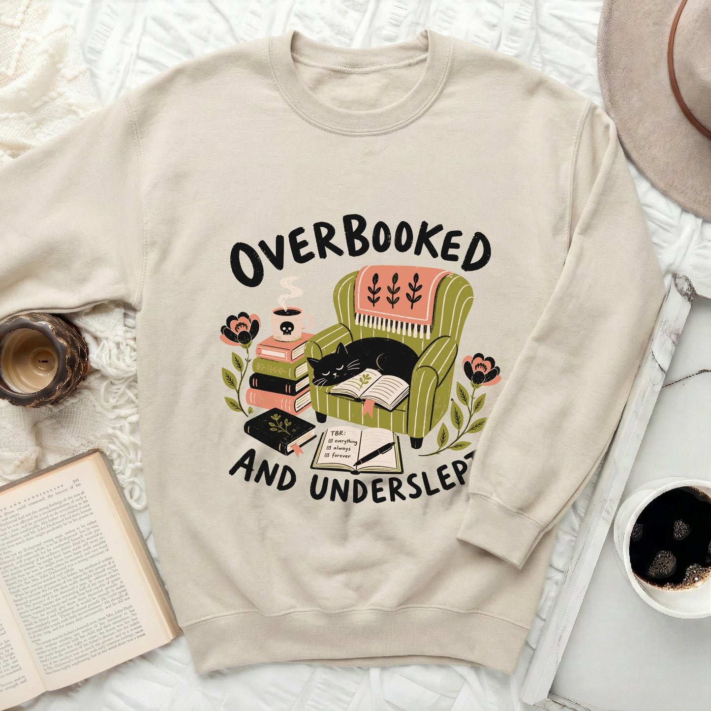 Cream sweatshirt with "Overbooked and Underslep" graphic featuring a cat reading in a chair.