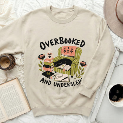 Cream sweatshirt with "Overbooked and Underslep" graphic featuring a cat reading in a chair.
