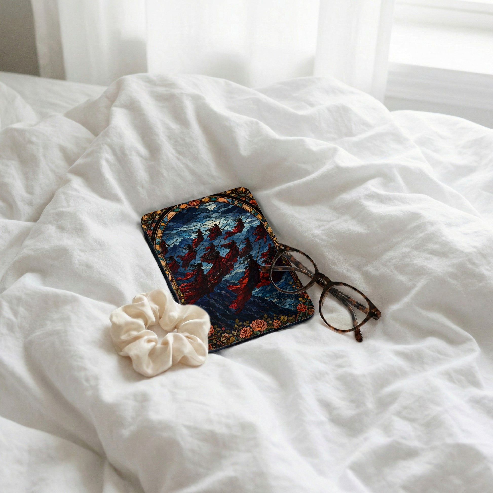 Mousepad with stained glass design of flowing figures rests on white bedding with scrunchie and glasses.