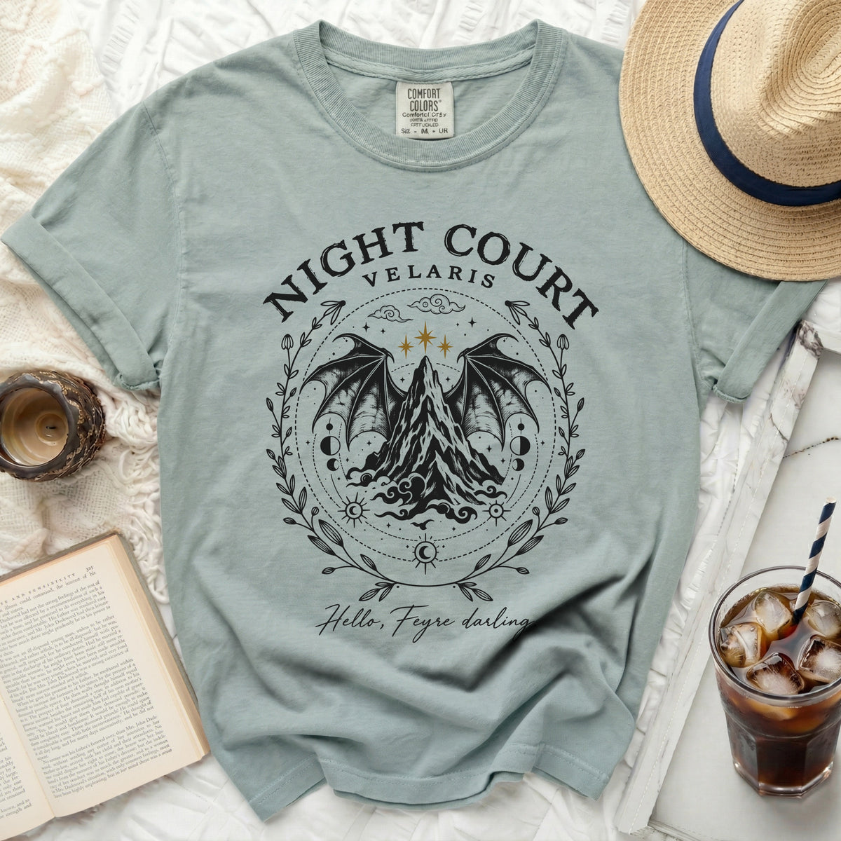 A sage green t-shirt with a black graphic of mountains, wings, and Night Court Velaris text.