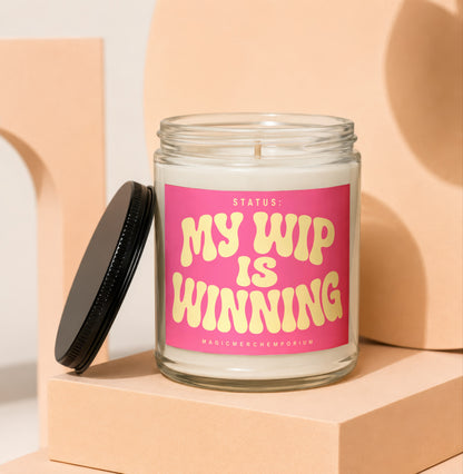 Glass candle with pink label reading "STATUS: MY WIP IS WINNING"