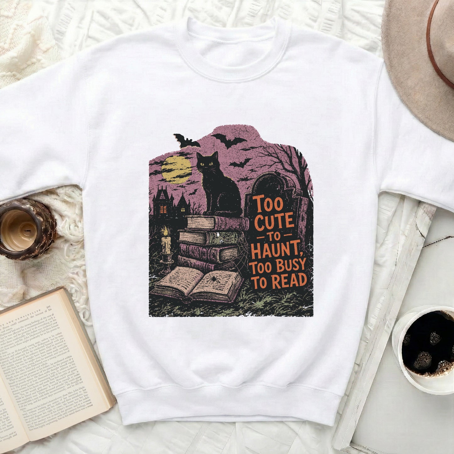 White sweatshirt with Halloween graphic of black cat, books, haunted house, bats, and moon.