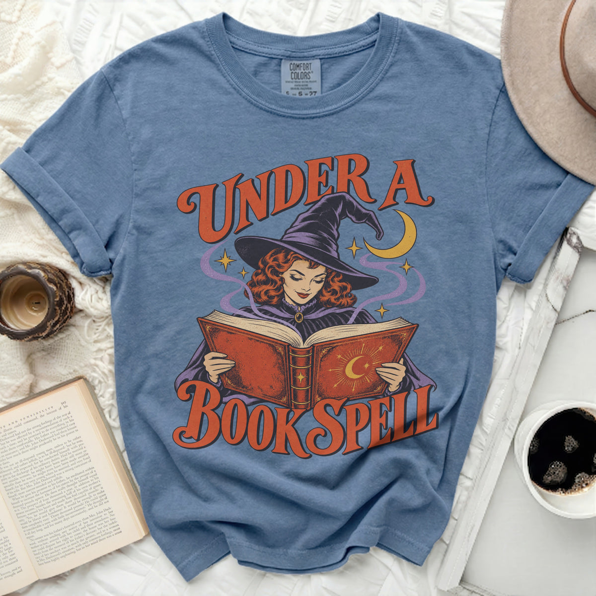 Blue t-shirt with graphic of witch reading under crescent moon and stars.