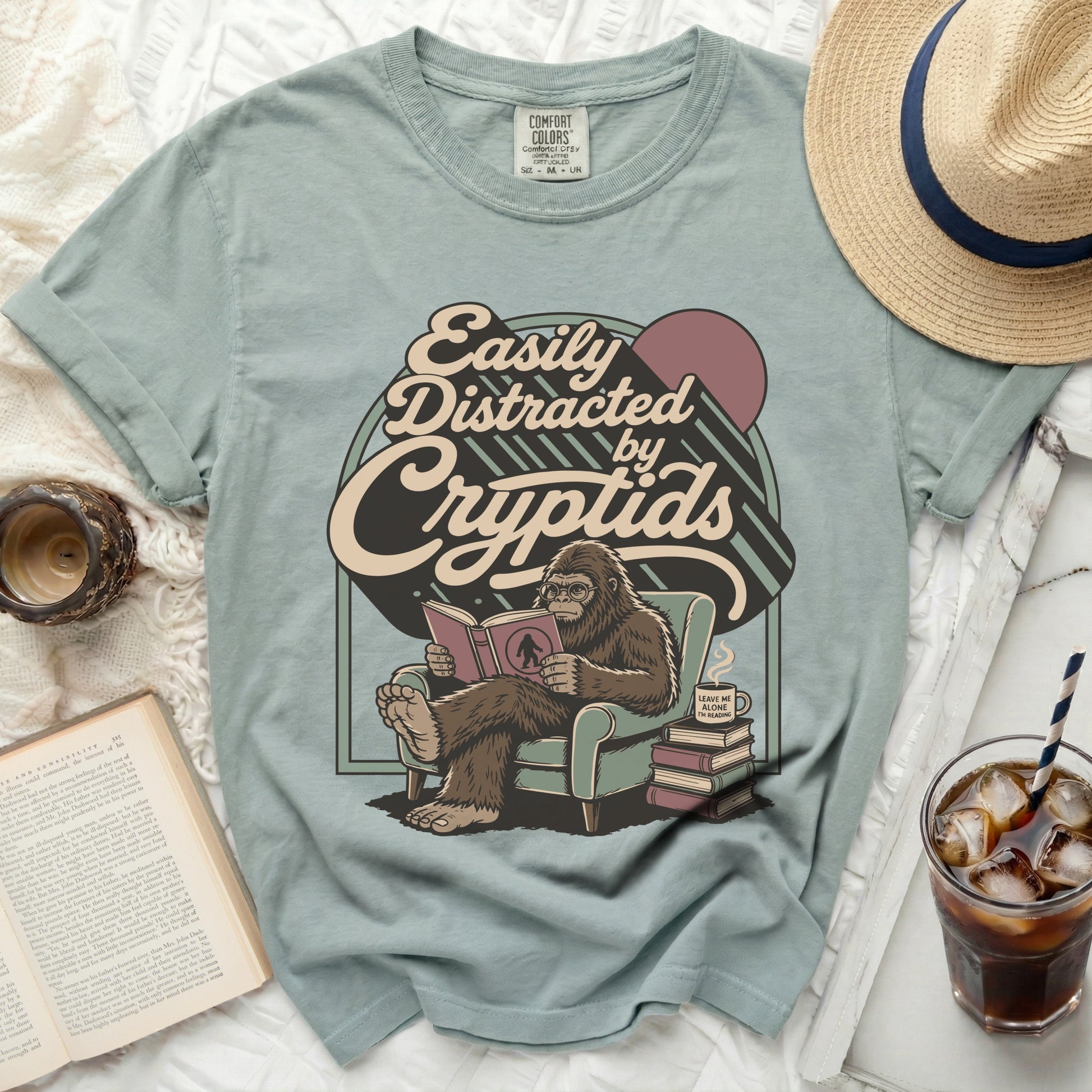 Bigfoot sitting in a chair reading a book "Easily Distracted by Cryptids".