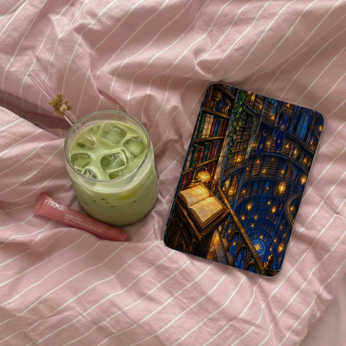 A glass of matcha latte, a lip balm, and an open book with a fantasy library scene.