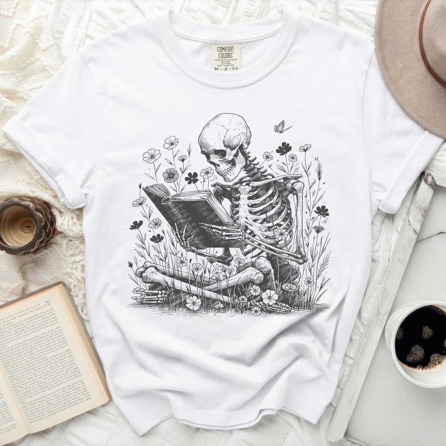 White t-shirt with a skeletal figure reading a book in a floral setting.