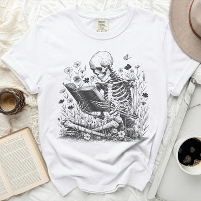 White t-shirt with a skeletal figure reading a book in a floral setting.