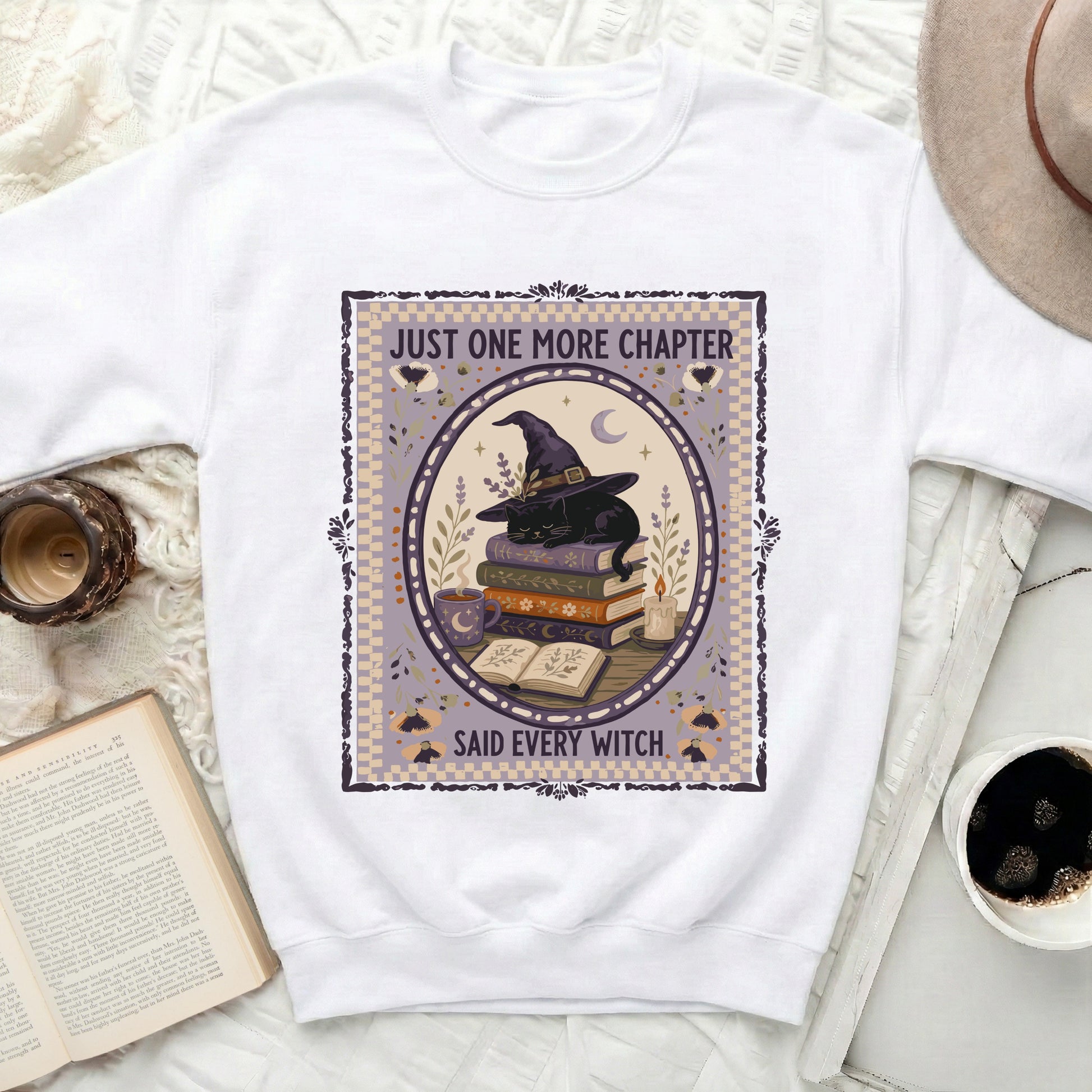 White sweatshirt with black cat, books, hat, and text "Just One More Chapter Said Every Witch".