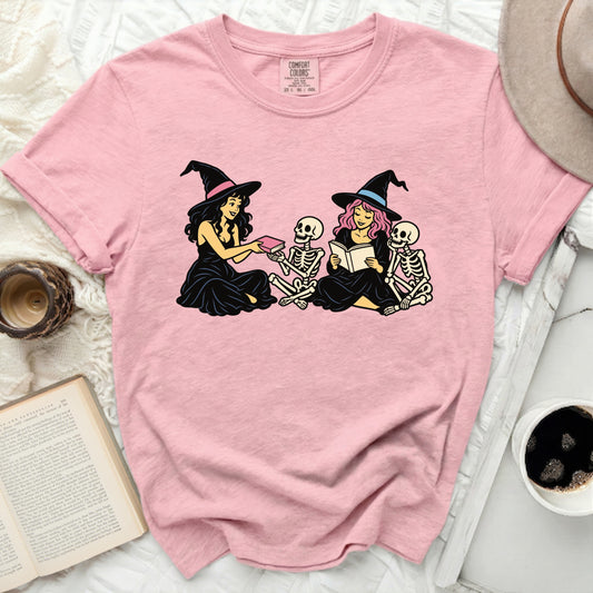 Pink t-shirt with illustration of two witches and skeletons reading books.