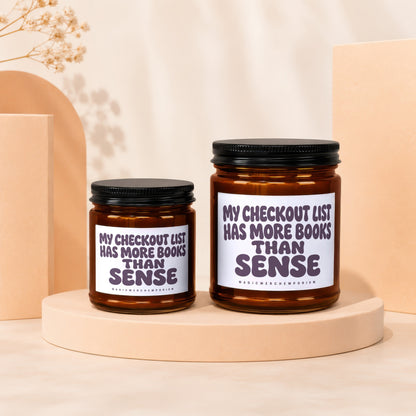 Two amber glass candles with black lids display white labels with purple text: "My checkout list has more books than sense".