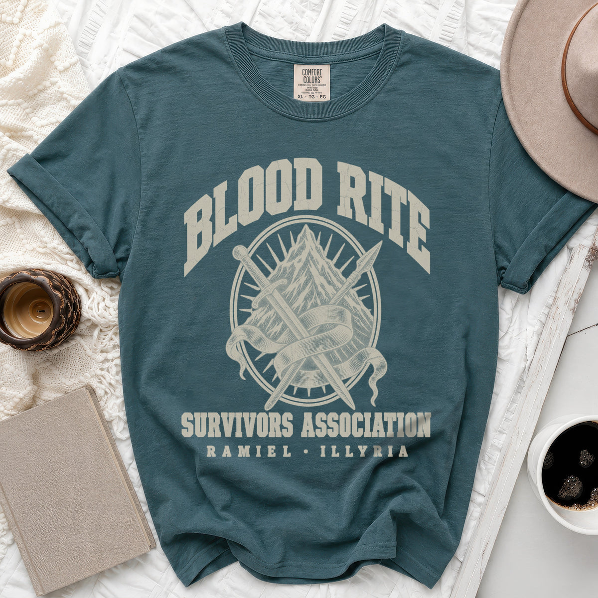A blue-green t-shirt with a graphic of crossed swords, a mountain, and the text Blood Rite Survivors Association.