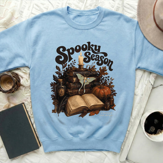 Light blue sweatshirt with "Spooky Season" print featuring books, candle, moth, and pumpkin.