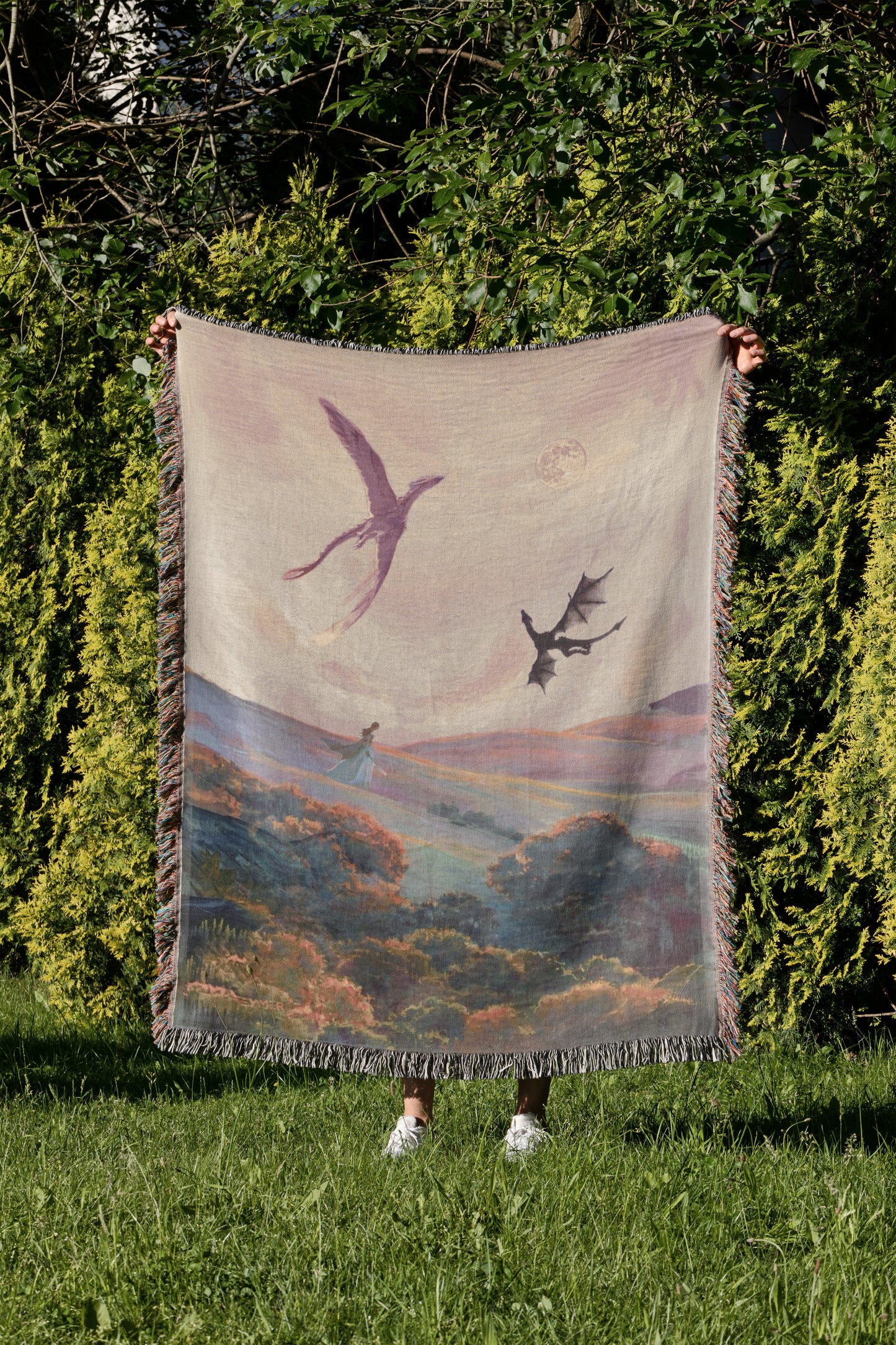 A person is standing in a grassy area, holding up a large blanket with a painting of a bird and a dragon on it.