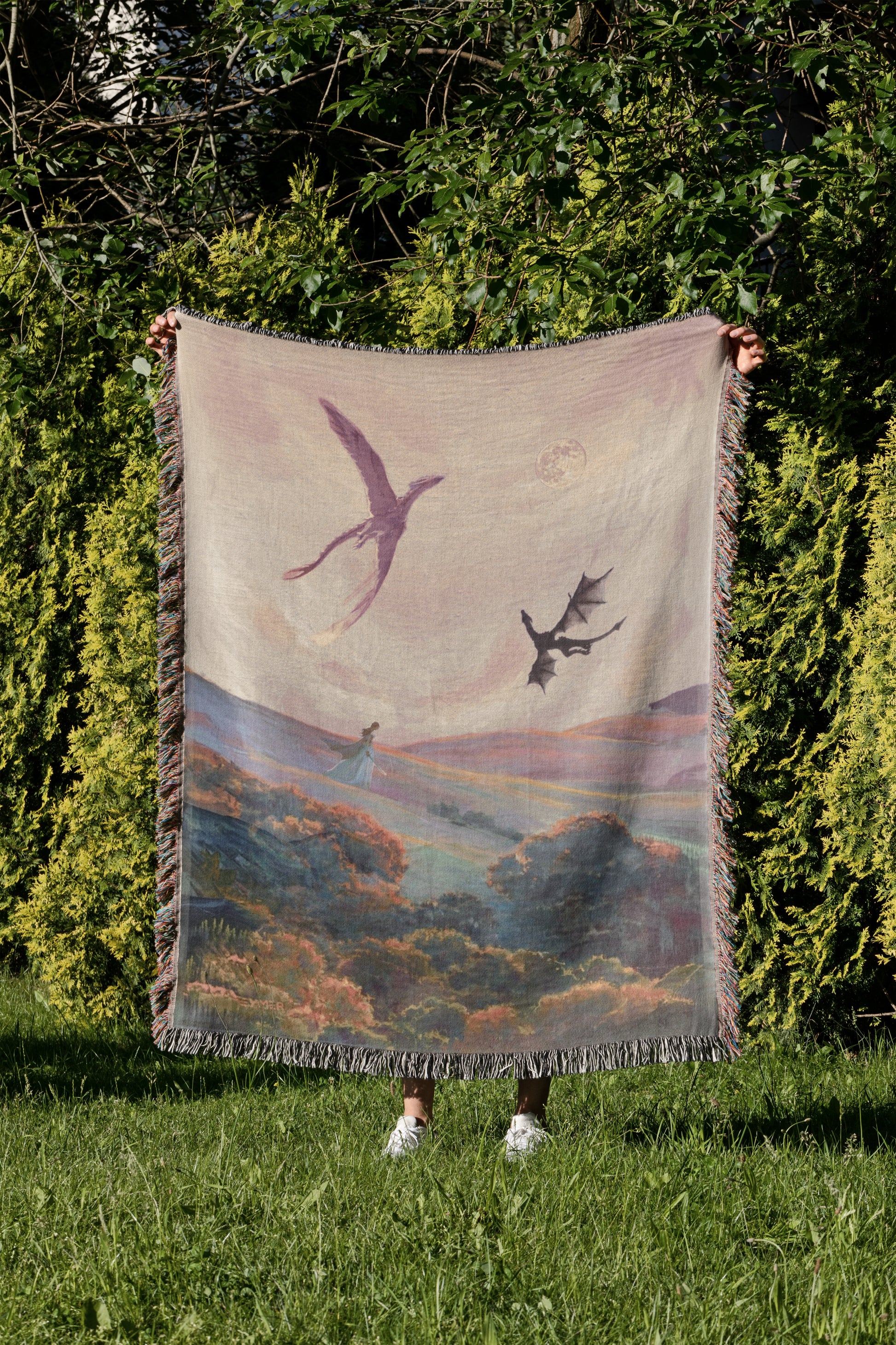 A person is standing in a grassy area, holding up a large blanket with a painting of a bird and a dragon on it.