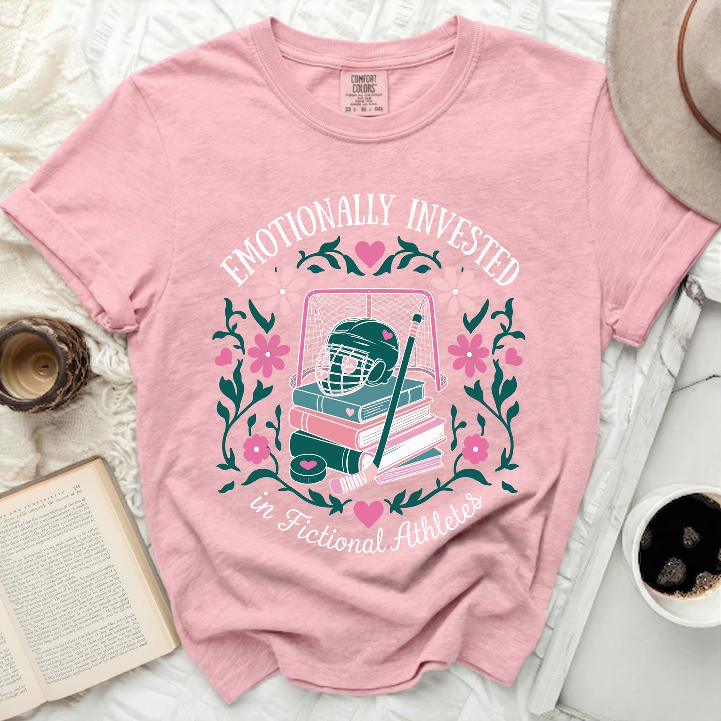Pink t-shirt with hockey gear, books, and floral design, text "Emotionally Invested in Fictional Athletes."