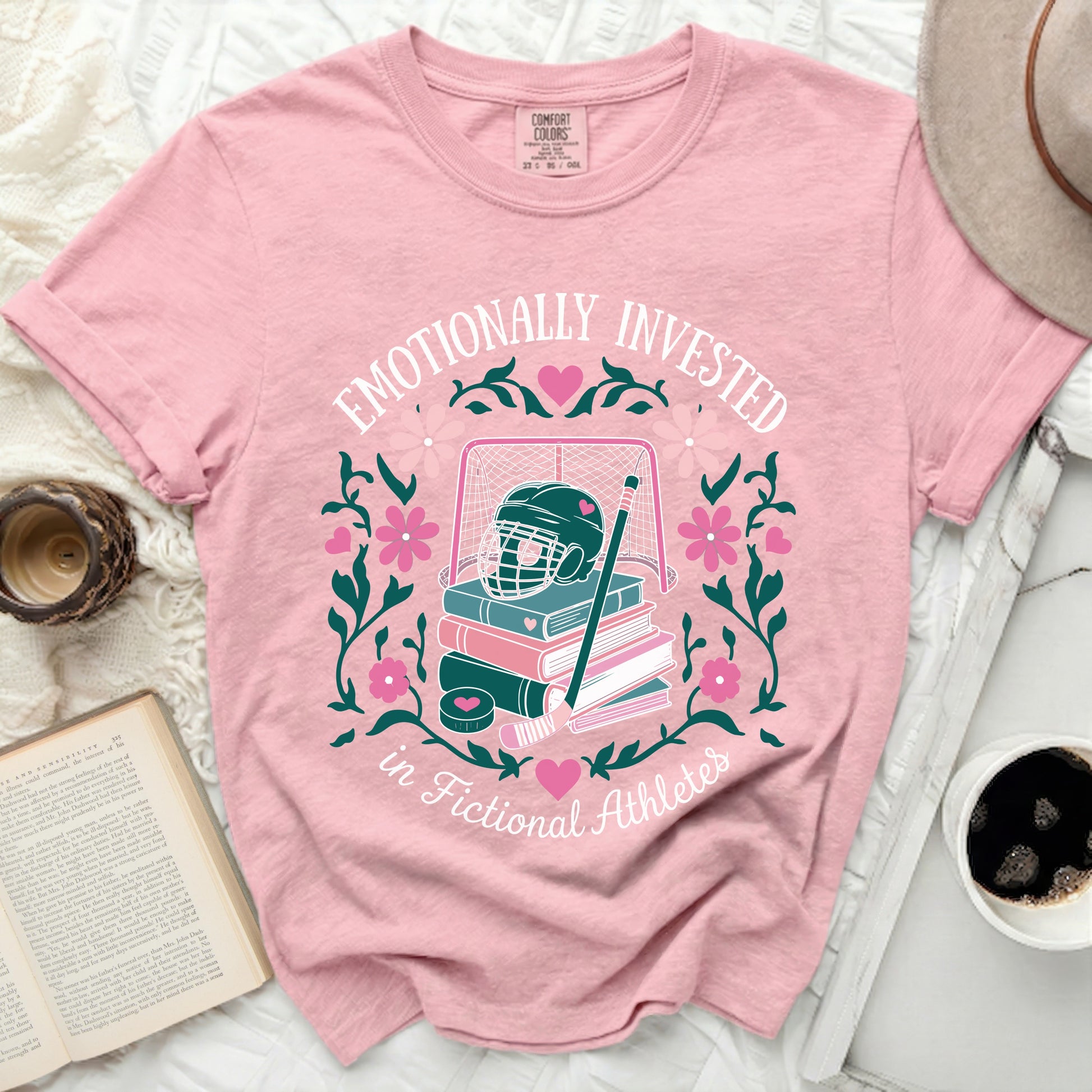 Pink t-shirt with hockey gear, books, and floral design, text "Emotionally Invested in Fictional Athletes."